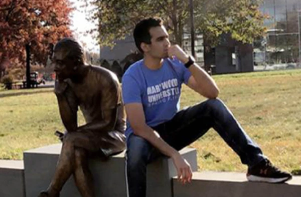 A young man in a blue t-shirt and jeans sitting next to a bronze statue of a seated figure, mimicking the statue's pose with a hand under his chin.